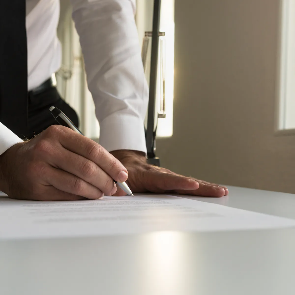 A person signing a document at a desk in window light