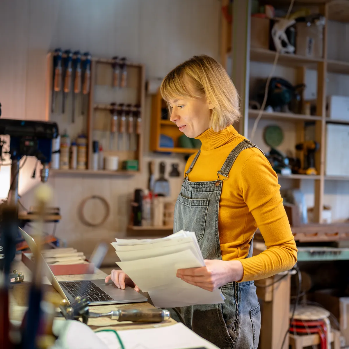 A workshop owner reading a stack of paperwork beside her laptop